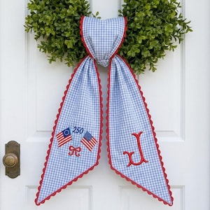 May include: A light blue and white gingham fabric door hanger with red scalloped edges. One side features two embroidered American flags with the number 250, while the other side displays a large red embroidered letter J in a script font.