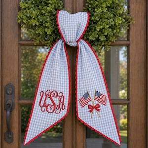 May include: A patriotic door scarf with a blue and white gingham pattern, red scalloped edges, and embroidered details. Features the letters "USA" in red script and two American flags with a red bow. Perfect for Independence Day or other patriotic holidays.