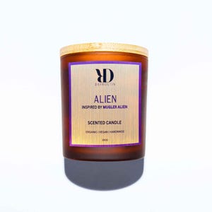 Può includere: Una candela profumata in vetro marrone con coperchio in legno. L'etichetta presenta la parola "ALIEN" e il testo "INSPIRED BY MUGLER ALIEN". La candela è etichettata "SCENTED CANDLE" e "ORGANIC | VEGAN | HANDMADE". La candela pesa 300g.
