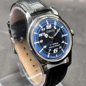 May include: An Oris wristwatch with a black leather strap, a silver-tone metal case, and a round dial featuring a black center, a blue outer ring, white Arabic numerals, silver hands, and the text Oris, 17 Jewels, Shock Proof, and Swiss Movt.