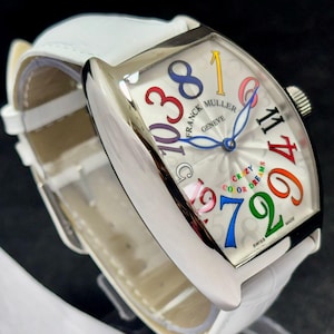 Könnte beinhalten: Eine luxuriöse Armbanduhr mit weißem Lederarmband und silberfarbenem Gehäuse. Das Zifferblatt ist weiß mit bunten Ziffern und den Worten "CRAZY COLOR DREAMS". Die Uhr ist mit "SWISS MADE" gekennzeichnet.