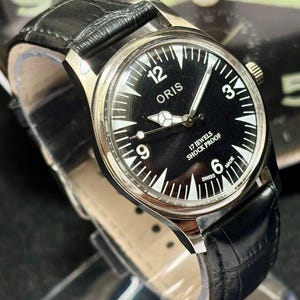 Puede incluir: Un reloj de pulsera ORIS vintage con esfera negra y marcadores blancos. El reloj presenta una caja plateada y una correa de cuero negro. La esfera incluye el texto "ORIS", "17 JEWELS", "SHOCK PROOF" y "SWISS MADE".