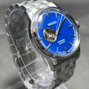 May include: A Seiko Presage Automatic watch with a vibrant blue sunray dial, silver-tone hands, and hour markers. The watch features an open-heart aperture at the nine o'clock position, a polished stainless steel case, and a matching link bracelet.