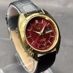 Puede incluir: Un reloj de pulsera automático Seiko 5 con esfera redonda de color rojo intenso, marcadores de hora y manecillas doradas. El reloj tiene una caja dorada pulida y una correa de cuero negro con acabado texturizado. La esfera muestra el texto Seiko, Automatic, 17 Jewels y Japan Made.