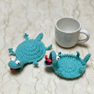 Könnte beinhalten: Zwei türkisfarbene Häkeluntersetzer in Monsterform mit weißen Augen und roten Zungen, neben einer weißen Keramiktasse. Die handgefertigten Untersetzer haben ein skurriles, verspieltes Design und verleihen jedem Tisch eine lustige Note.
