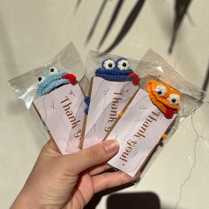 May include: Three crocheted frog-like ornaments in clear plastic packaging. Each frog has large eyes and a red tongue. The frogs are blue, orange, and blue. Each package has a brown tag with the words "Thank you!" printed on it.