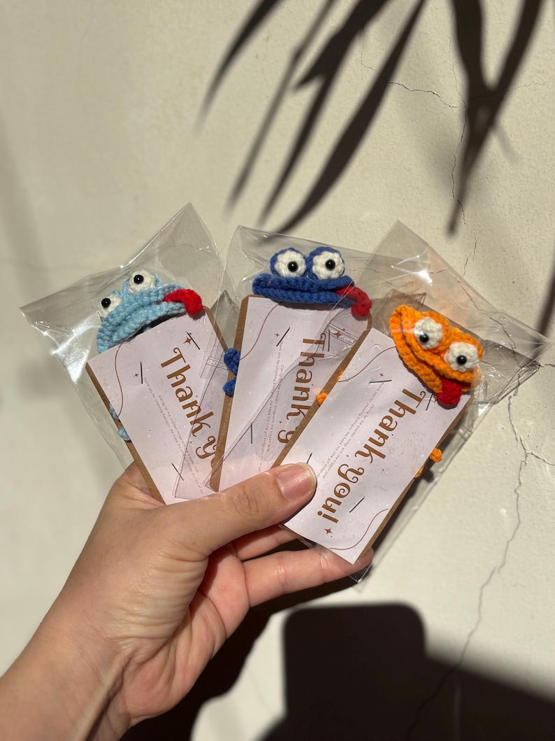 May include: Three crocheted frog-like ornaments in clear plastic packaging. Each frog has large eyes and a red tongue. The frogs are blue, orange, and blue. Each package has a brown tag with the words "Thank you!" printed on it.