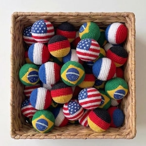 Puede incluir: Una cesta de mimbre llena de bolas de ganchillo, cada una adornada con las banderas de diferentes países. Las bolas presentan las banderas de Estados Unidos, Alemania, Francia y Brasil. Las bolas son rojas, blancas, azules, amarillas y verdes.