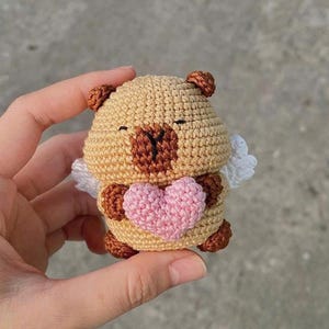 May include: A crocheted bear figurine in tan, brown, and pink. The bear has small white wings and is holding a pink heart. The bear's ears, nose, and feet are brown. The figurine is handmade.