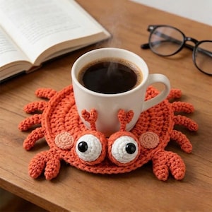 May include: A white coffee mug filled with dark coffee sits on an orange crocheted crab coaster. The coaster has large, googly eyes and crab claws. A book and glasses are in the background.