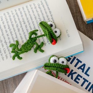 May include: Two handmade green crochet alligator bookmarks with white eyes and red tongues rest on open books. One bookmark is on the page, and the other is on top of the book. The books have text in Vietnamese.