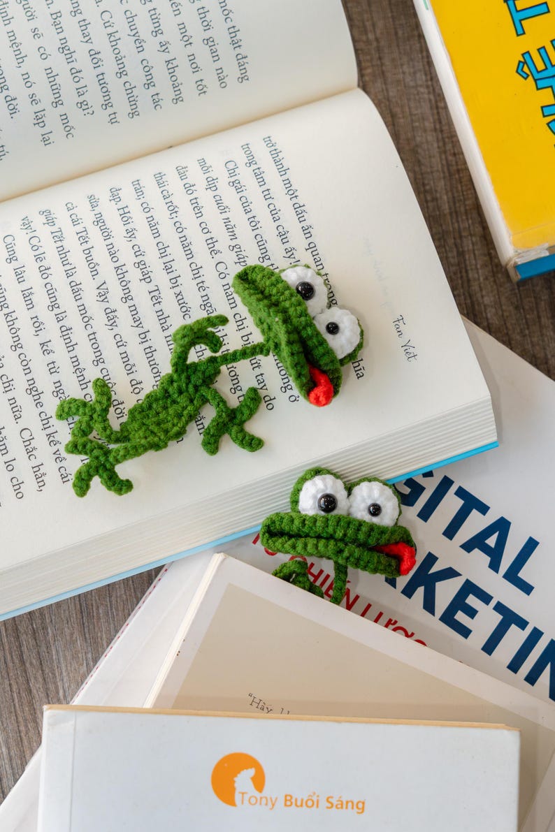 May include: Two handmade green crochet alligator bookmarks with white eyes and red tongues rest on open books. One bookmark is on the page, and the other is on top of the book. The books have text in Vietnamese.