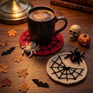 May include: A dark brown ceramic mug sits on a round red crocheted coaster with a black spiderweb pattern and a small beige crocheted spider. Nearby is a beige crocheted coaster with a black spiderweb and a black spider, a skull, and a pumpkin.