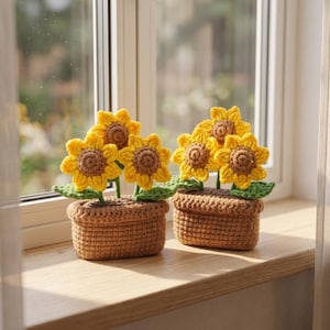May include: Two crochet sunflower arrangements in brown pots sit on a wooden windowsill. Each arrangement features bright yellow sunflowers with brown centers and green leaves. The pots are a light brown color.