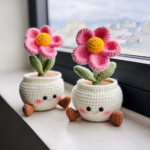 May include: Two handmade crochet flower pots with smiling faces. Each pot has a pink flower with a yellow centre, green leaves, and brown feet. The pots are white and sit on a windowsill.