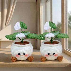Puede incluir: Dos macetas de ganchillo hechas a mano con caras sonrientes y pequeños pies marrones. Cada maceta blanca contiene una planta de ganchillo verde y blanca. Las plantas tienen hojas grandes y detalladas. Las macetas están sobre un alféizar de madera.