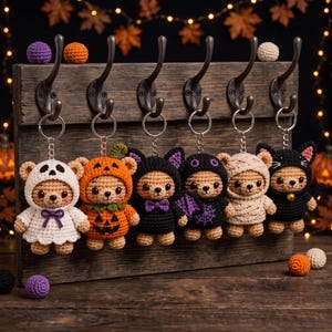 Op de afbeelding: Zes gehaakte beren-sleutelhangers hangend aan een houten rek. De beren dragen Halloween-kostuums: een spook, een pompoen, een vleermuis, een outfit met spinnenwebpatroon, een mummie en een zwarte kat met een klein belletje.