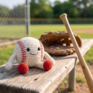 May include: A crocheted baseball with a smiling face, red stitching, and red feet sits on a wooden bench. A baseball glove and a wooden baseball bat lean against the bench in the background, suggesting a baseball theme.