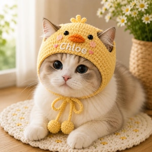 May include: A yellow crocheted pet hat featuring a chick design with an orange beak, black eyes, and small pink heart accents. The hat includes the name Chloe embroidered in white thread and two dangling yellow pom-poms attached to chin ties.