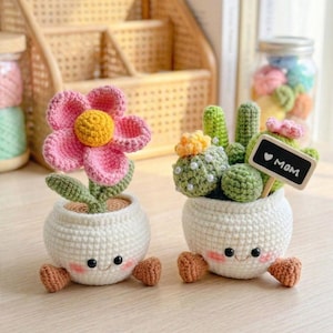 Könnte beinhalten: Zwei gehäkelte Blumentöpfe mit lächelnden Gesichtern. Einer hat eine rosa Blume mit gelber Mitte, der andere grüne Kaktusformen und ein kleines Schild mit der Aufschrift "MOM". Beide Töpfe haben braune Füße.