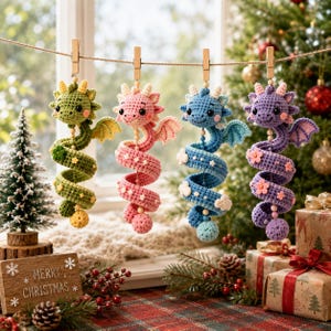 May include: Four crocheted dragon ornaments in green, pink, blue, and purple, each featuring a coiled body, small wings, and decorative floral accents. The dragons are suspended from a string by wooden clothespins in a festive holiday setting.