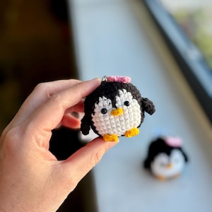 Puede incluir: Un llavero de pingüino tejido a mano. El pingüino es negro y blanco con pies amarillos y un lazo rosa. Tiene un anillo plateado para el llavero. Otro pingüino es visible en el fondo, también con un lazo rosa.