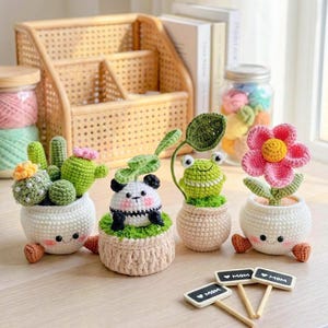 May include: A collection of handmade crochet plant pot dolls, including a cactus, panda, frog, and flower. Each doll is in a white pot with smiling faces and small brown feet. Small signs with "MOM" are in front of the dolls.