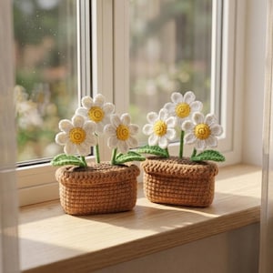 May include: Two crochet daisy flower arrangements in brown pots sit on a wooden windowsill. Each pot contains several white daisies with yellow centers and green leaves. The window is in the background.