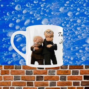 May include: White ceramic mug with a handle, featuring two Muppet characters in suits. The mug has text on the right side. The background is a blue gradient with water droplets, and the bottom is a brick wall.