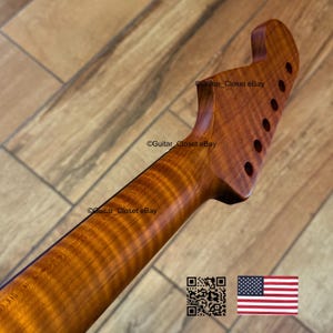 May include: A close-up of a guitar neck and headstock made of figured wood. The wood grain has a prominent flame pattern with a warm, amber color. The headstock has six tuning peg holes. The image includes a QR code and an American flag.