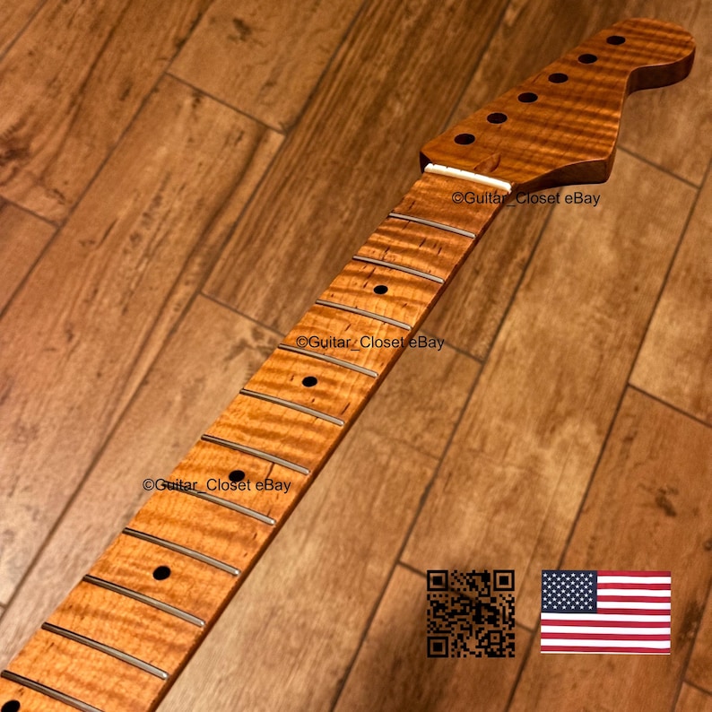 May include: A guitar neck featuring a flamed maple wood grain pattern with a natural amber finish. The neck includes a fretboard with metal frets, dark circular position markers, and a headstock with six pre-drilled holes for tuning pegs.