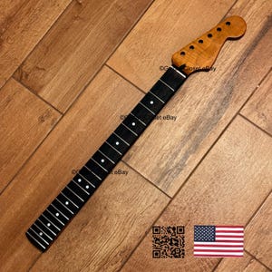 May include: A guitar neck with a natural wood finish on the headstock and a dark fretboard. The headstock has tuning peg holes. The fretboard has white dot inlays. The neck is angled on a wooden surface.