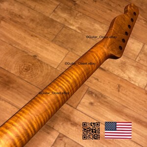 May include: A guitar neck with a flamed maple finish. The neck has a headstock with tuning peg holes and a smooth, polished surface. The wood grain is visible, with a warm, amber color. The background is a wooden surface.