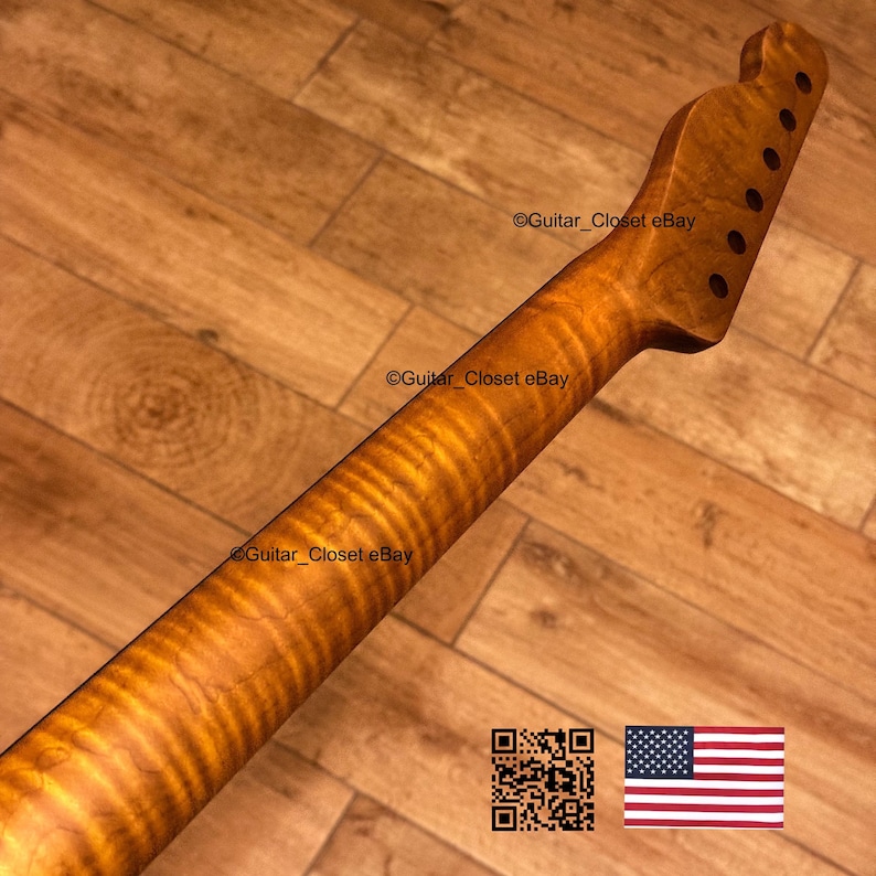 May include: A guitar neck with a flamed maple finish. The neck has a headstock with tuning peg holes and a smooth, polished surface. The wood grain is visible, with a warm, amber color. The background is a wooden surface.