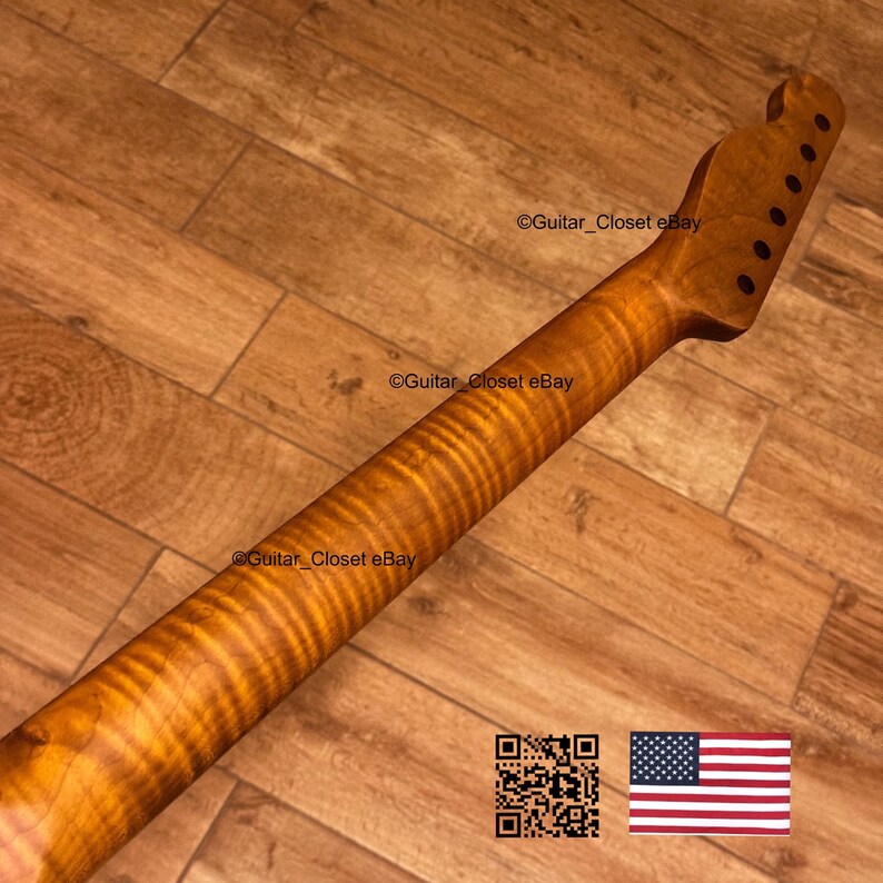 May include: A close-up of a guitar neck with a flamed maple finish. The wood grain has a wavy pattern with shades of brown and amber. The headstock has pre-drilled holes for tuning pegs.
