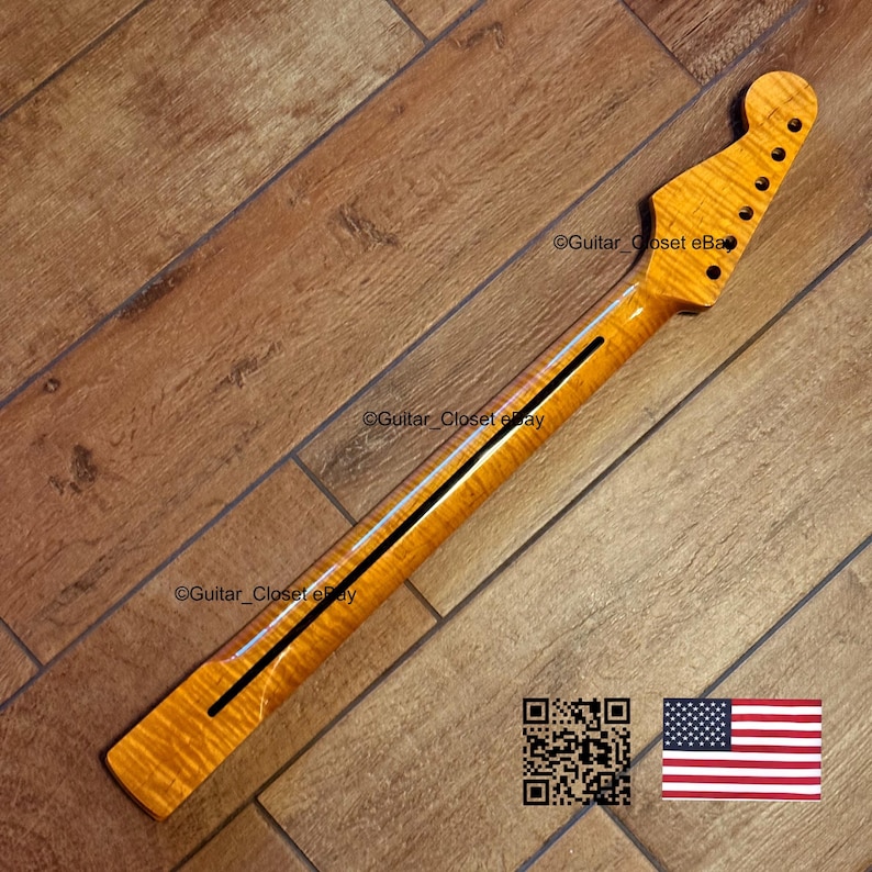 May include: A guitar neck with a flamed maple finish. The neck has a dark stripe down the center and a headstock with tuning peg holes. The wood grain is visible. The background is a wooden floor.
