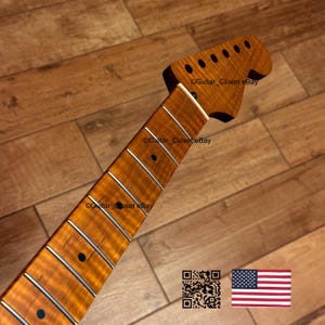 May include: A guitar neck with a flamed maple finish. The neck has a light brown colour with dark brown streaks. The fretboard has silver frets and black dot inlays. The headstock has six tuning peg holes. The image also includes a QR code and an American flag.