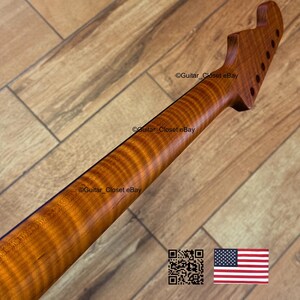 May include: A close-up of a guitar neck made of figured maple wood. The neck has a natural wood finish with a wavy grain pattern. The headstock has pre-drilled holes for tuning pegs. The image includes a QR code and the American flag.