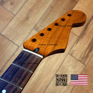 May include: A guitar neck with a natural wood finish. The headstock has six tuning peg holes and a dark fretboard with silver frets. The wood grain is visible, and the neck is angled. The image includes a QR code and a small American flag.