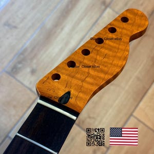 May include: A guitar neck with a flamed maple headstock and a dark fretboard. The headstock has six tuning peg holes. The wood has a warm, amber color. The fretboard has silver frets. The image includes a QR code and an American flag.