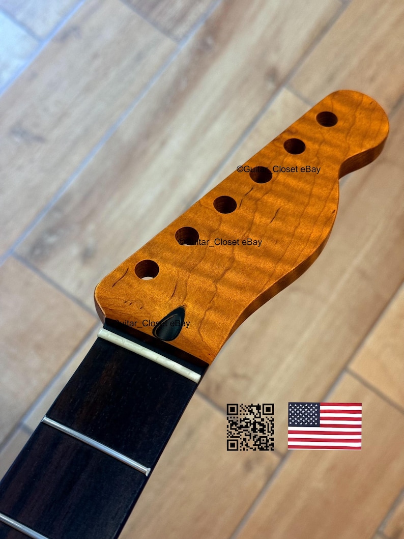 May include: A guitar neck with a flamed maple headstock and a dark fretboard. The headstock has six tuning peg holes. The wood has a warm, amber color. The fretboard has silver frets. The image includes a QR code and an American flag.