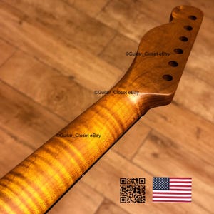 May include: A close-up of a guitar neck with a flamed maple finish. The neck has a natural wood grain pattern with a warm, amber hue. The headstock features tuning peg holes. The image includes a QR code and an American flag.