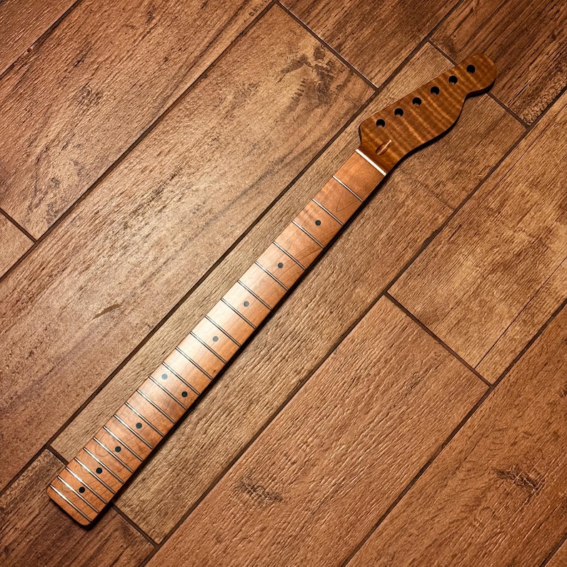 Op de afbeelding: Een gitaarhals met een lichtgekleurde toets, donkere ronde positieaanduidingen en metalen frets. De kop is gemaakt van gevlamd, amberkleurig hout met zes voorgeboorde gaten voor stemmechanieken, gerangschikt in een enkele rij.