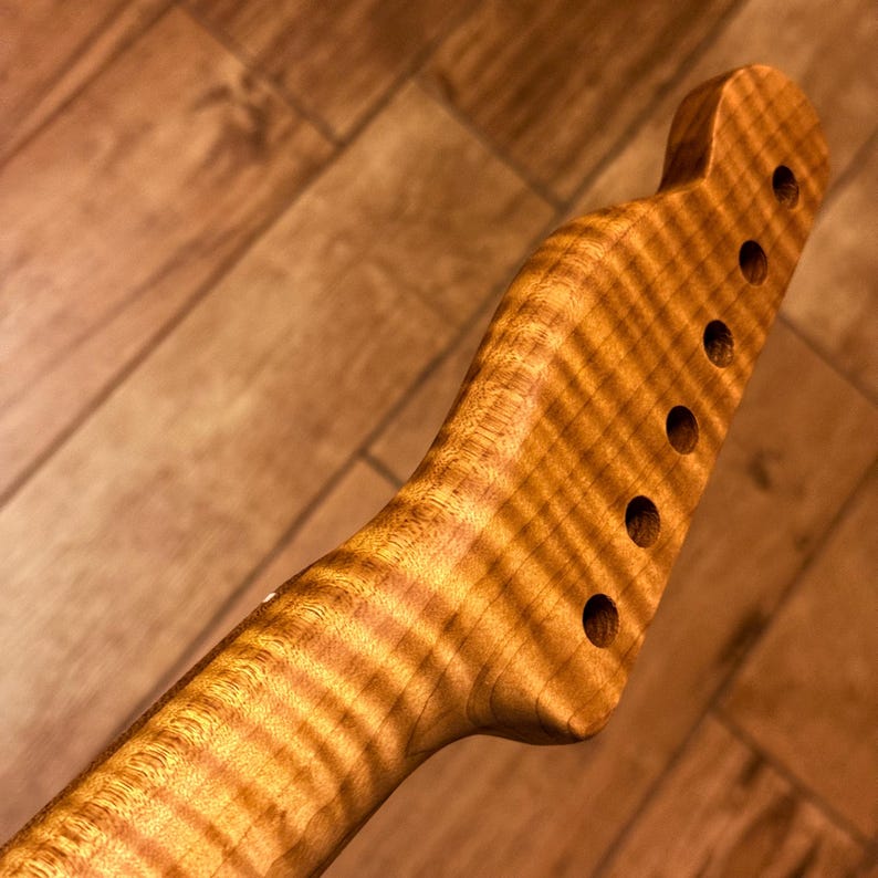 Op de afbeelding: Een gitaarhals met een kop voorzien van zes geboorde gaten voor stemmechanieken. Het hout vertoont een opvallend vlamachtig esdoornnerfpatroon met een warme, goudbruine afwerking. De hals loopt vloeiend over in de vorm van de kop.