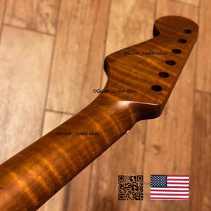 Op de afbeelding: Een close-up van een gitaarhals van gevlamd hout. De hals heeft een warme bruine kleur met een glanzende afwerking. De kop heeft zes gaten voor de stemknoppen. De tekst "Guitar Closet eBay" is zichtbaar.