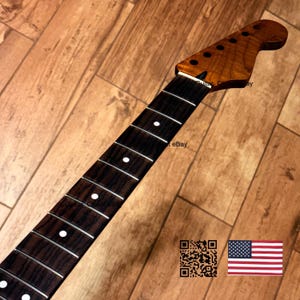 May include: A guitar neck featuring a dark brown wood fretboard with white circular position markers and silver frets. The headstock is made of a light brown wood with six circular holes for tuning pegs. A QR code and a US flag are visible.