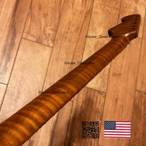 Op de afbeelding: Een close-up van een gitaarhals met een gevlamde esdoorn afwerking. De hals heeft een warme bruine kleur met een gestreept patroon. De kop heeft gaten voor de stemknoppen. De tekst "Guitar_Closet eBay" is zichtbaar.