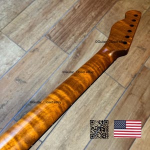 May include: A close-up of a guitar neck with a flamed maple finish. The neck is a warm, amber color with dark brown grain patterns. The headstock has tuning peg holes. The image includes a QR code and a small American flag.