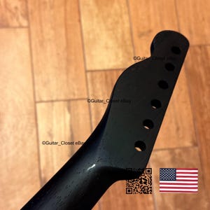 May include: A black guitar neck with six tuning peg holes. The headstock has a rounded shape. The neck is angled and has a smooth, dark finish. The image includes a QR code and a small American flag.