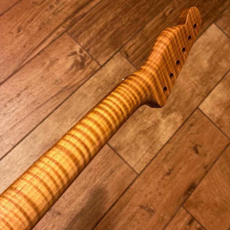 Op de afbeelding: Een gitaarhals gemaakt van lichtbruin hout met een opvallend tijgerstreep vlam-esdoorn nerfpatroon. De kop is schuin geplaatst en bevat zes cirkelvormige gaten op een rij voor stemmechanieken, met een gladde, gepolijste afwerking.